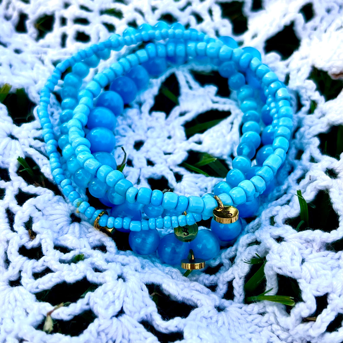 Close-up of the Coastal Blue Bliss bracelet stack on white crochet, showing blue beads and small gold-tone signature charms.