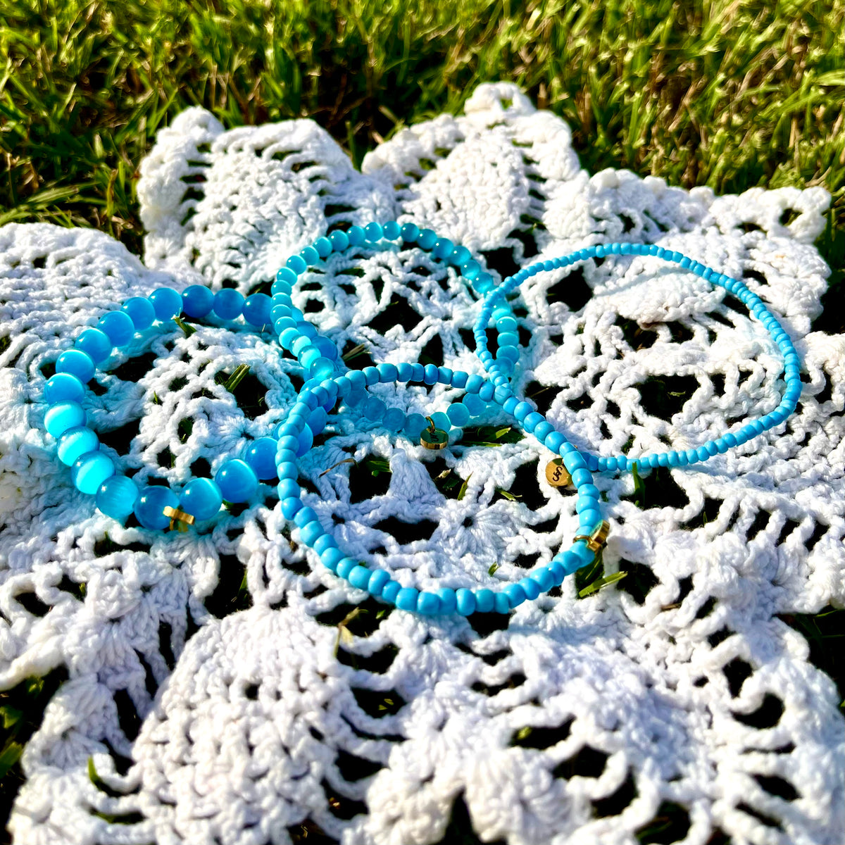 Coastal Blue Bliss bracelets spread across white crochet on grass, showing blue beads in varied sizes and gold-tone charms.